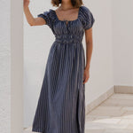 Woman in a striped dress standing against a white wall with greenery.