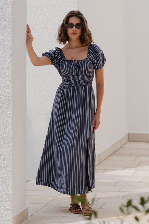 Woman in a striped dress standing against a white wall with greenery.