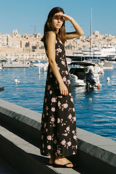 Woman in a floral dress standing by a waterfront with boats and buildings in the background