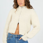 Woman wearing a cream zip-up sweater and blue jeans on a white background