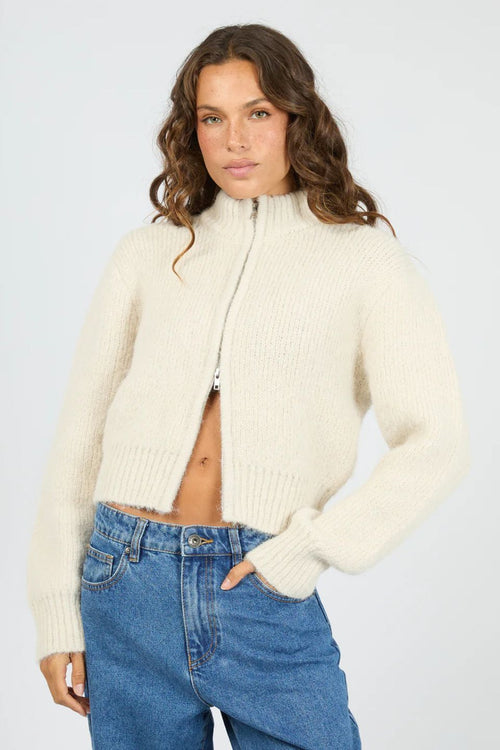 Woman wearing a cream zip-up sweater and blue jeans on a white background