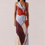 A woman modeling a halter-neck maxi dress with an abstract geometric print in red, light blue, cream, and burgundy with gold lines, set against a solid light beige background.
