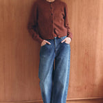 Person wearing a brown cardigan and blue jeans standing against a wooden wall.