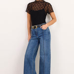 A model stands against a plain white studio background wearing a black short-sleeve top with a sheer lace upper section, blue wide-leg jeans with a gold belt, and black sandals.