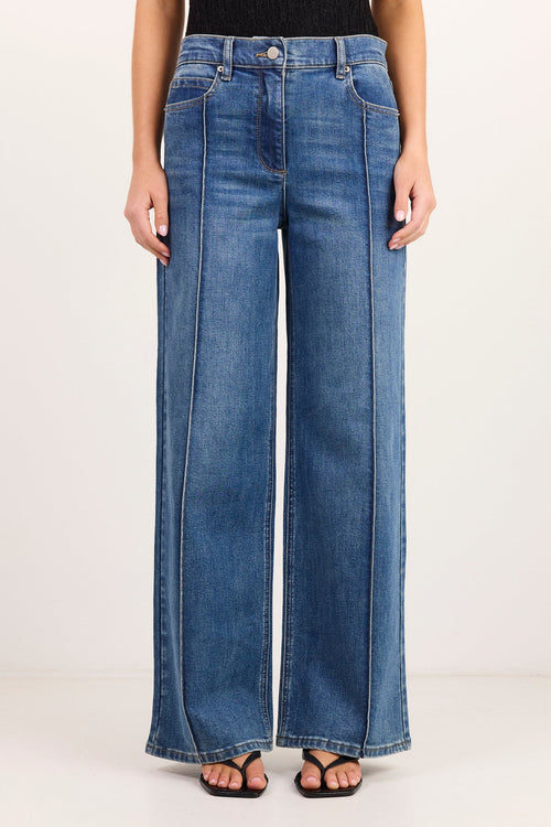 A model from the waist down wearing blue wide-leg denim jeans, a black top, and black sandals, standing against a plain white studio background.