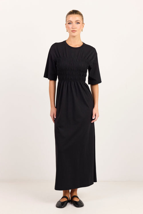 A woman modeling a black midi dress with short sleeves, a round neckline, and a smocked bodice, standing against a plain white studio background.