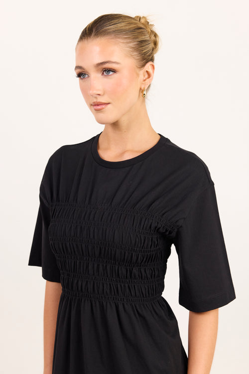 A woman wearing a black short-sleeve dress with a smocked bodice, photographed against a plain light background.