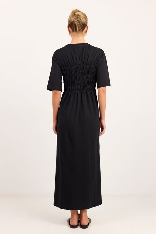 A woman viewed from the back wearing a black midi dress with short sleeves and a smocked bodice, standing against a plain white studio background.