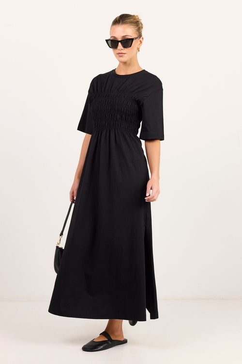 A woman modeling a black midi dress with short sleeves, a round neckline, and a smocked waist, styled with black sunglasses, black flats, and a small black handbag against a plain white studio background.