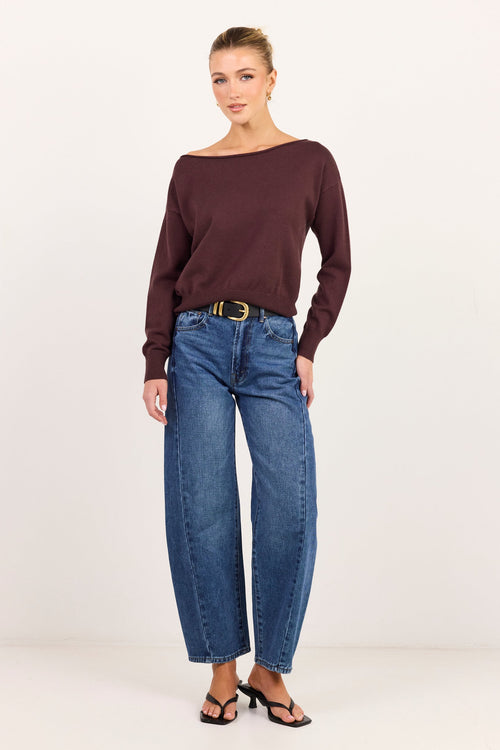 A woman modeling a dark burgundy off-the-shoulder long-sleeve knit top with blue wide-leg jeans and black sandals, standing against a plain white studio background.