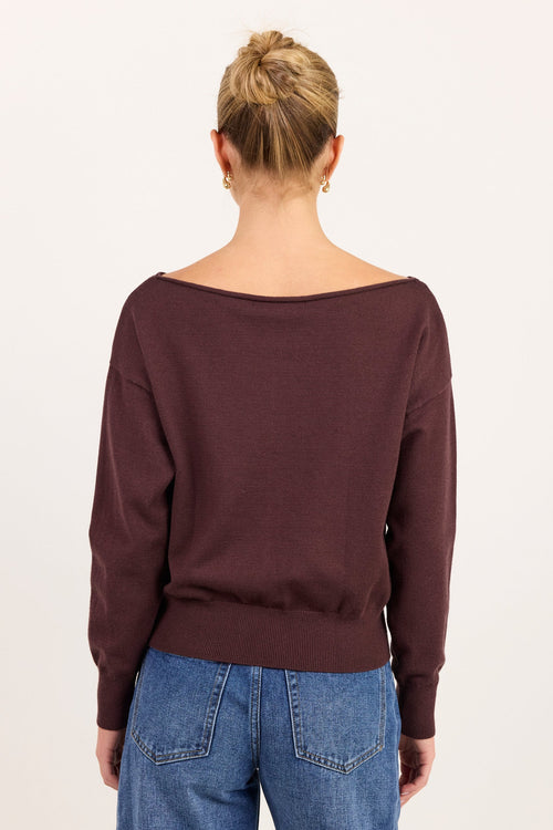 A woman viewed from the back wearing a dark plum-brown wide-neck sweater and blue jeans against a plain off-white background.