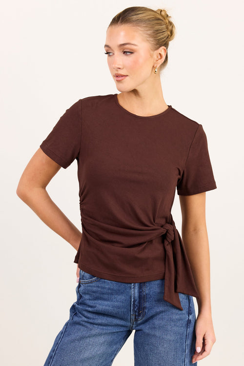 A woman modeling a short-sleeve brown knit top with a side twist detail, paired with blue jeans, against a plain off-white background.