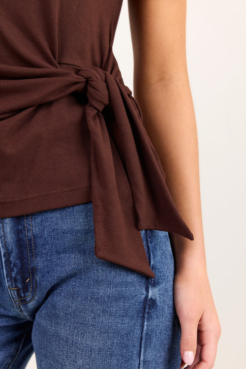 Close-up of a person wearing a dark brown sleeveless top tied at the waist, paired with blue jeans, against a plain light background.