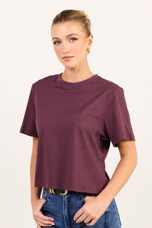 A woman modeling a short-sleeve crewneck t-shirt in a deep plum purple color, paired with blue jeans, against a plain off-white studio background.