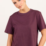 A woman wearing a short-sleeve crewneck t-shirt in a deep plum purple color, paired with blue jeans, against a plain off-white background.