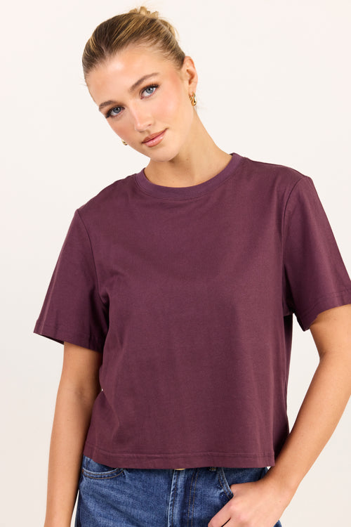 A woman wearing a short-sleeve crewneck t-shirt in a deep plum purple color, paired with blue jeans, against a plain off-white background.
