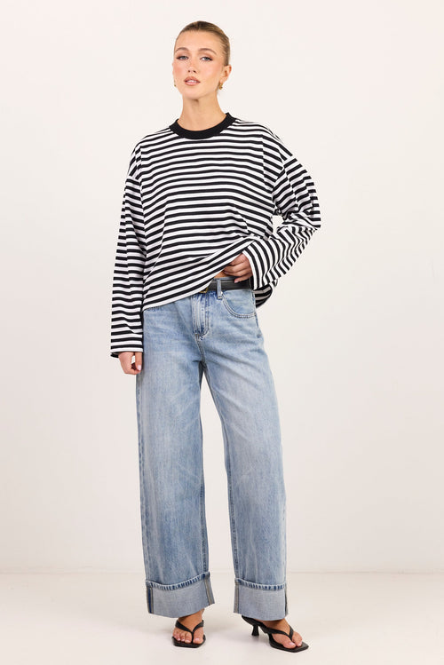 A model stands against a plain off-white studio background wearing a black and white horizontally striped long-sleeve top, light-wash wide-leg blue jeans, and dark brown heeled sandals.