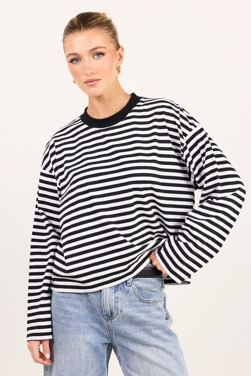 A woman modeling a black and white horizontally striped long-sleeve top with a black crew neck, paired with light-wash blue jeans, against a plain off-white background.