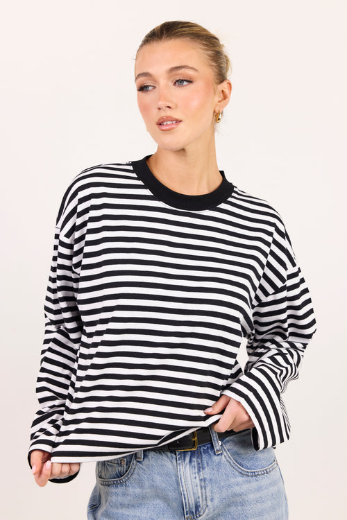 A woman wearing a black and white horizontally striped long-sleeve top with a black crew neck, paired with light blue jeans, against a plain light background.