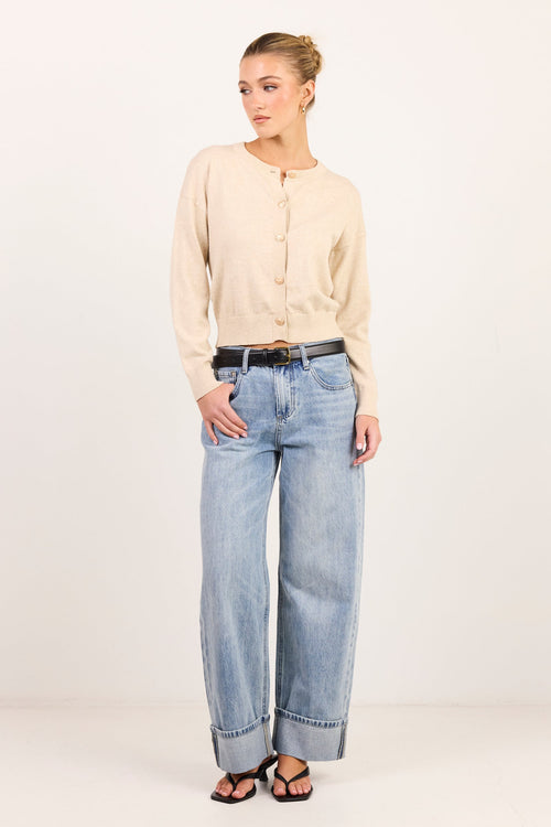 A woman modeling a light beige long-sleeve button-up shirt and light-wash wide-leg blue jeans with a black belt, standing against a plain white studio background.