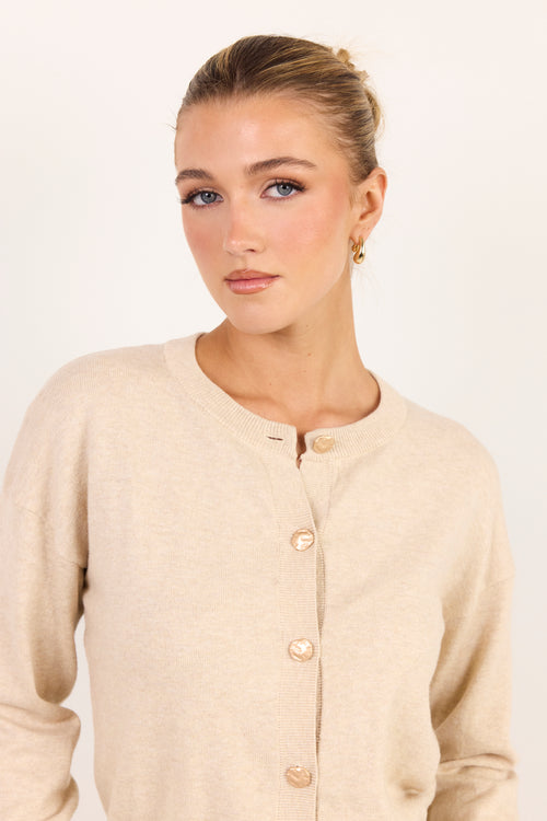 A woman wearing a light beige knit cardigan sweater with gold buttons, photographed against a plain off-white studio background.