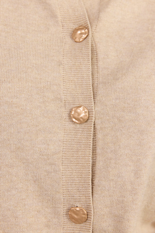 Close-up of a light beige knit cardigan with a vertical ribbed placket and four metallic gold buttons.