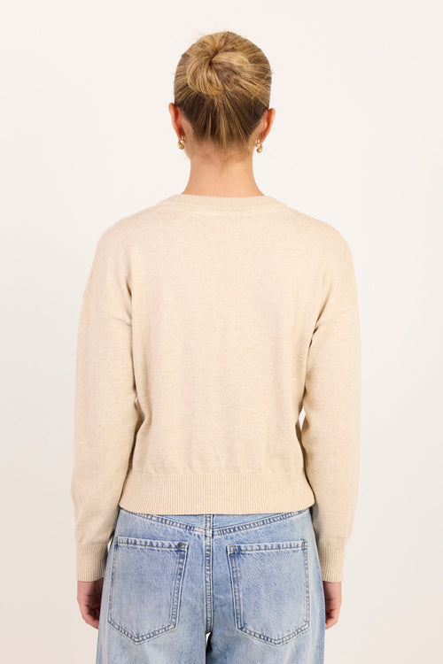 A woman viewed from behind wearing a light beige knit sweater and light blue jeans against a plain off-white studio background.