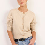 A woman modeling a beige knit cardigan with three-quarter sleeves and a button-up front, paired with light-wash blue jeans and a black belt, against a plain white studio background.