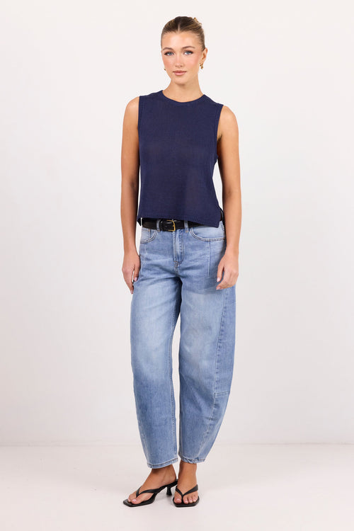 A woman modeling a sleeveless navy blue top, light-wash blue jeans, a black belt, and black sandals against a plain white studio background.