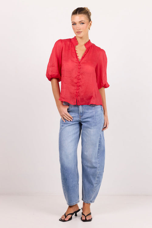 A woman modeling a red short-sleeve blouse with a ruffled V-neckline and front buttons, paired with light blue jeans and black sandals, standing against a plain white studio background.