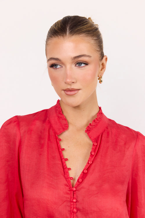 A woman wearing a red blouse with a ruffled high collar and decorative buttons down the front, photographed against a plain light background.