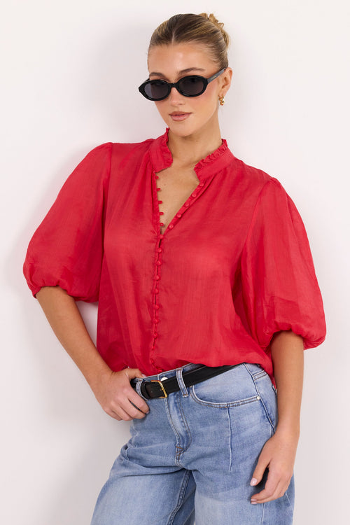 A woman modeling a red short-sleeve blouse with a ruffled collar and button front, paired with light blue jeans and black sunglasses, against a plain white background.