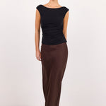 A model wearing a black cap-sleeve top and a dark brown satin midi skirt, styled with black sunglasses and black sandals, standing against a plain white studio background.
