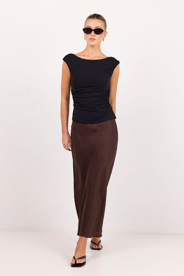 A model wearing a black cap-sleeve top and a dark brown satin midi skirt, styled with black sunglasses and black sandals, standing against a plain white studio background.