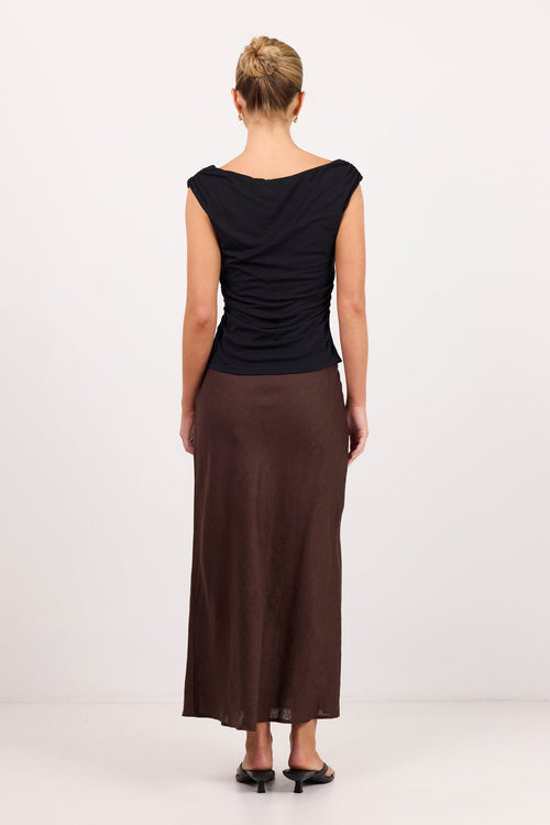 A woman viewed from the back wearing a black draped sleeveless top and a long brown skirt, standing against a plain light gray studio background.
