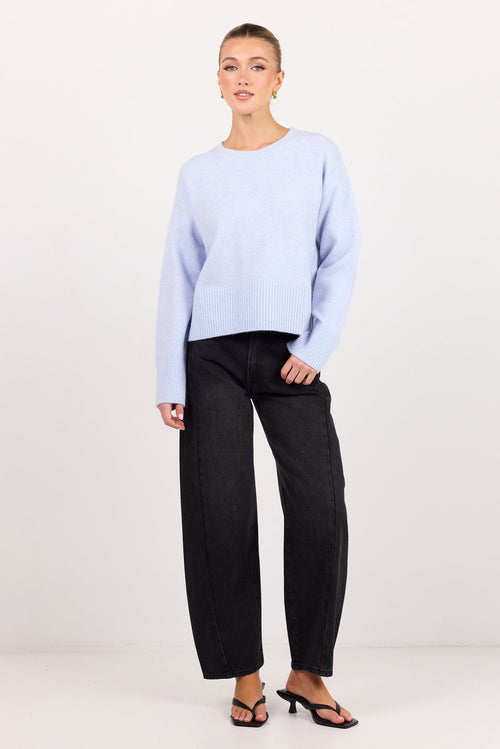 A model stands against a plain white studio background wearing a light periwinkle blue long-sleeve knit sweater, black wide-leg trousers, and black strappy mule sandals.