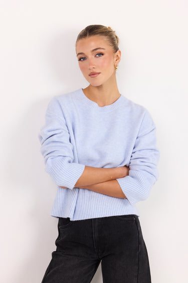 A woman wearing a light periwinkle blue ribbed knit sweater with black pants, standing against a plain white studio background.