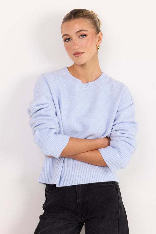 A woman wearing a light periwinkle blue ribbed knit sweater with black pants, standing against a plain white studio background.