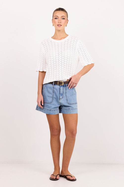 A woman modeling a white short-sleeve knit top and light blue denim shorts with a brown belt, standing against a plain white studio background.