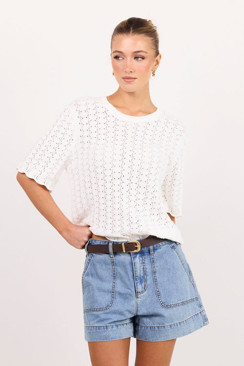 A woman modeling a white short-sleeve knit top and light blue denim shorts against a plain white studio background.