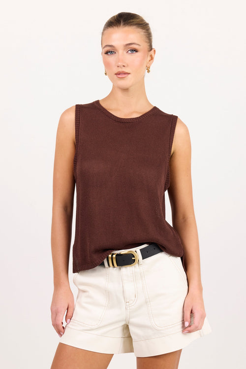 A woman modeling a sleeveless dark brown knit top paired with off-white shorts and a black belt with a gold buckle, set against a plain light gray studio background.