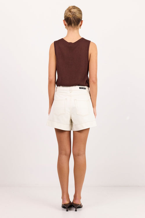 A woman viewed from behind wearing a sleeveless dark brown top and white shorts, standing against a plain white studio background.