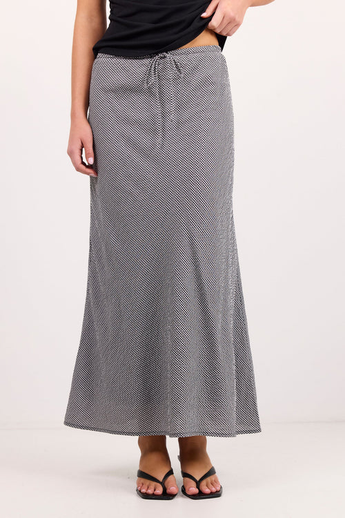 A woman modeling a long gray and white micro-patterned maxi skirt with a drawstring waist, paired with a black top and black sandals, against a plain white studio background.