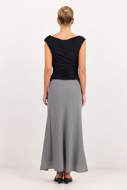 A woman viewed from behind wearing a black sleeveless top and a long gray skirt, standing against a plain light studio background.