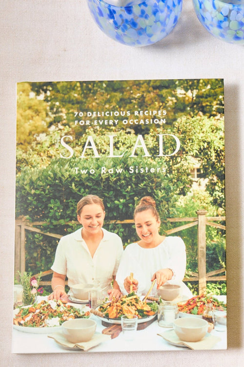 Cookbook titled 'Salad' by Two Raw Sisters on a table with decorative items.