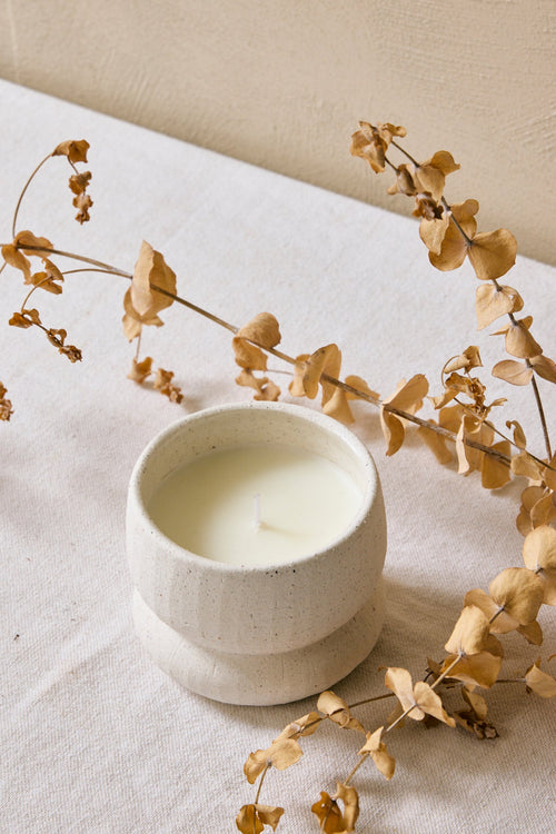 Ceramic candle holder with a lit candle on a textured surface with dried leaves.