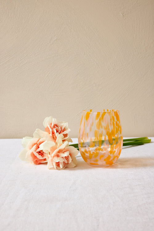 Glass with yellow and orange pattern next to pink flowers on a white surface