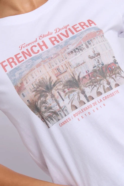 A close-up of a person wearing a white graphic T-shirt. The shirt features a large rectangular print of a French Riviera building and palm trees, with coral-red text reading 'FRENCH RIVIERA' and smaller text above and below.