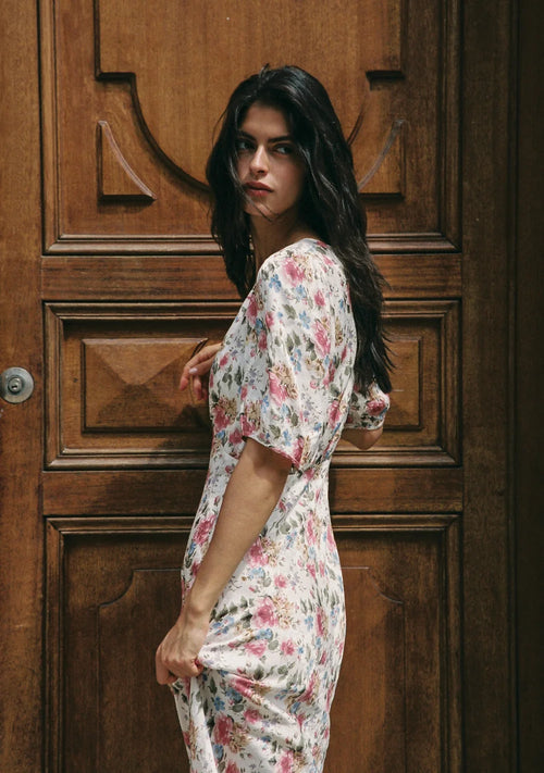 A woman with long dark hair stands in front of a carved wooden door, wearing a short-sleeve white dress with a pink and green floral print.