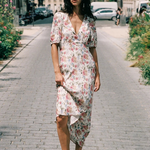 A woman wearing a short-sleeve floral midi dress and black slide sandals walks down a cobblestone city street lined with greenery and buildings.